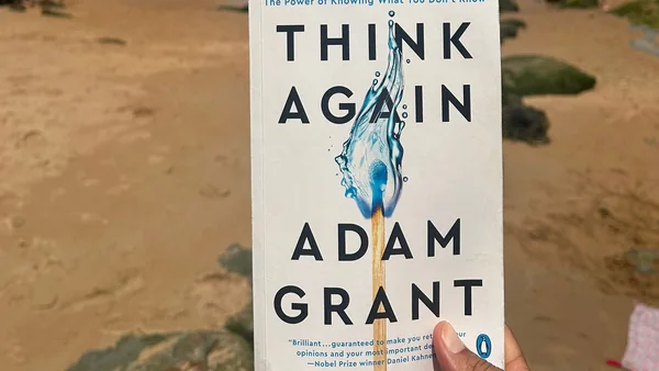 El libro Think Again, de Adam Grant, sostenido en una mano frente a una cala con la marea baja