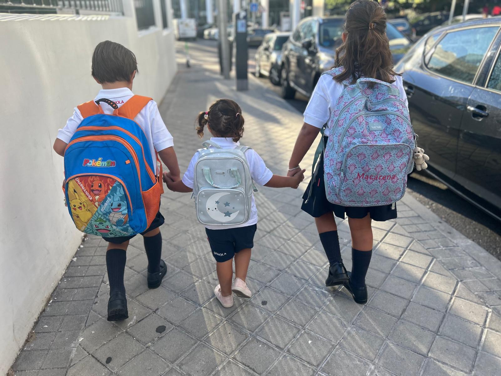 Three children in uniform with school backpacks walk hand in hand, seen from behind, toward school