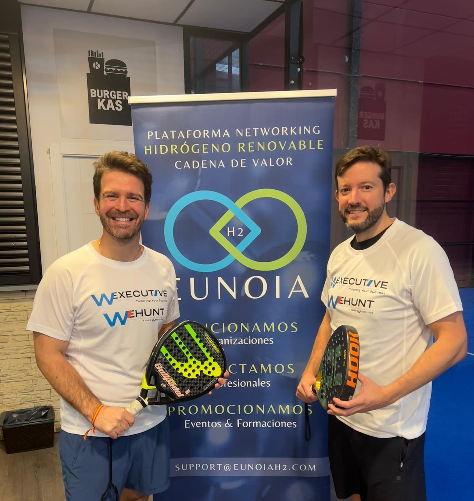 Two men holding padel rackets and wearing W Executive and WeHunt shirts in front of a Eunoia H2 roll-up