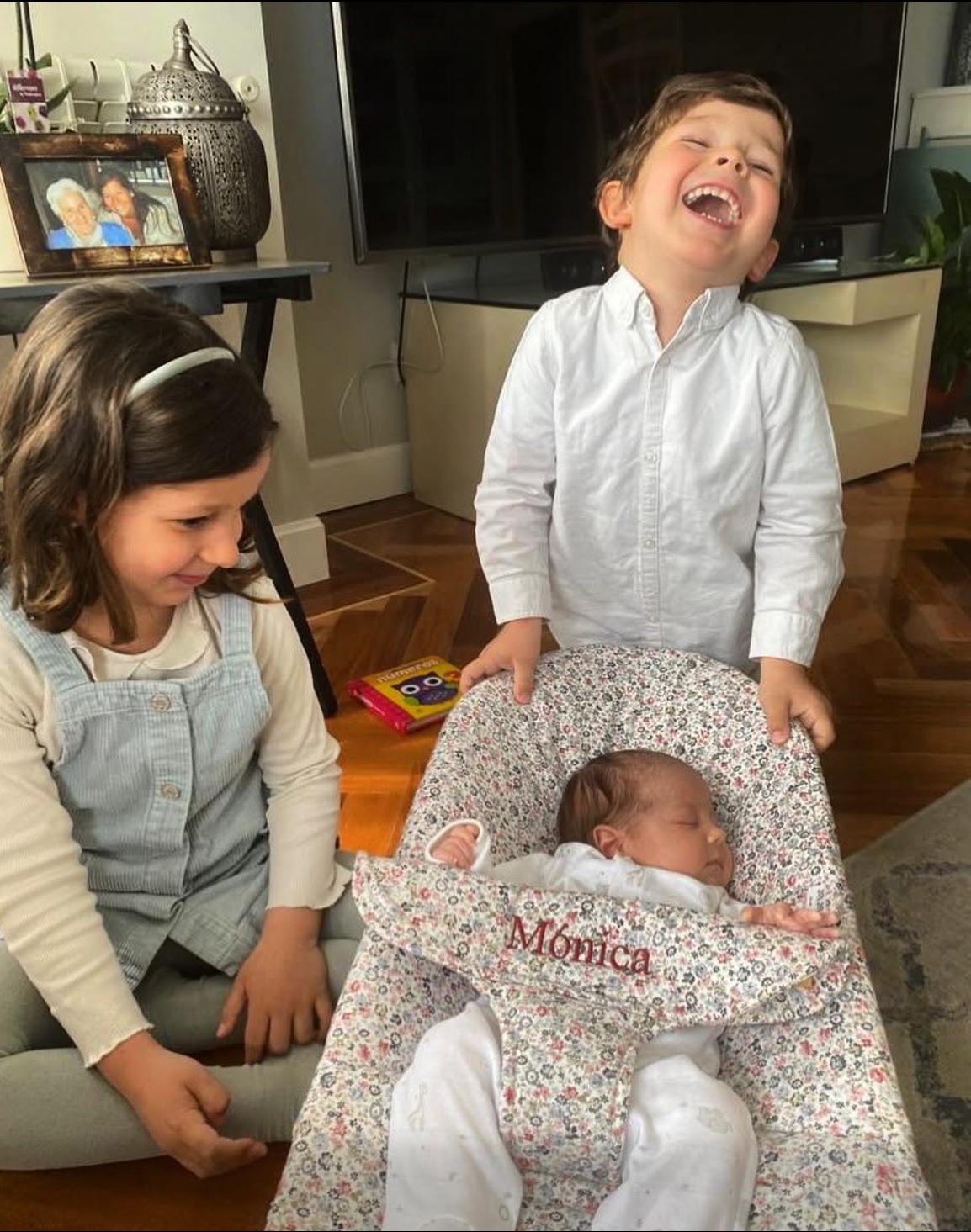 Two smiling kids next to their newborn sister in a bassinet with the name Mónica
